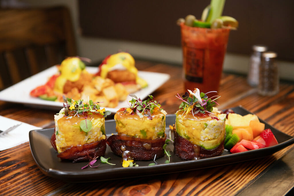 On a long black plate, three pieces of breakfast sushi are lovingly placed with a bloody mary in the back