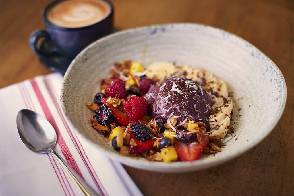 A bowl filled with blended acai with oats and fruits with coffee behind it
