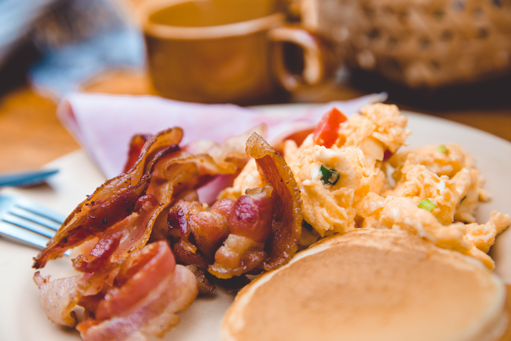 A pile of fresh bacon next to a stack of pancakes and a pile of eggs with coffee nearby