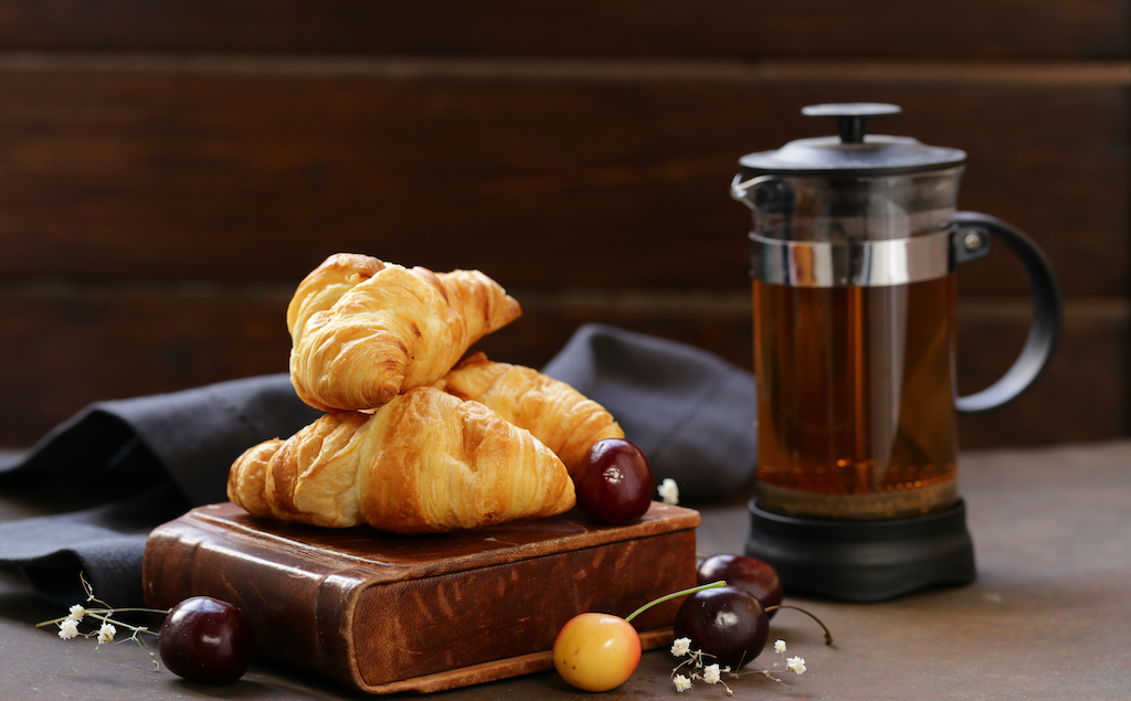 Three croissants on a wooden stand next to a pitcher of French press coffee