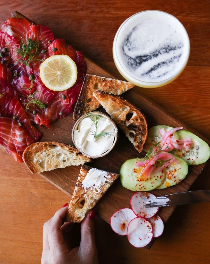 Precisely cut salmon with a lemon on top next to bread, radishes, cucumbers, and sauce