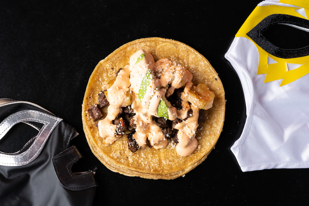 A little taco is humbly plated with seafood and meat, topped with sauces and garnish