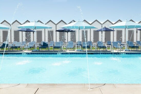 Pool in the front, then lounge chairs with umbrellas in the background, and small little huts with doors in the back