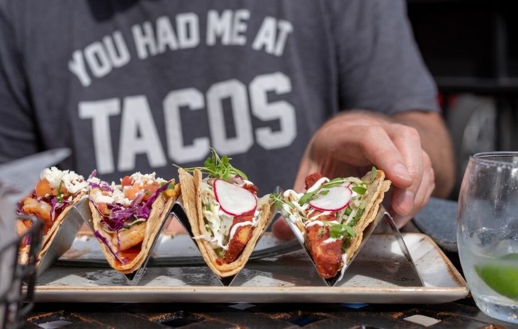 Man wearing grey tshirt that says: You had me at Tacos - in the foreground metal taco holder tray with 4 tacos