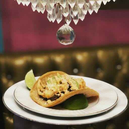 one taco on a stacked plate with a glass chandelier on top and a golden sofa and purple wall in the background