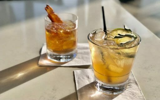 Two yellow/orange cocktails with ice. One with a slice of candied bacon, the other with a sprig of sage and lemon slice