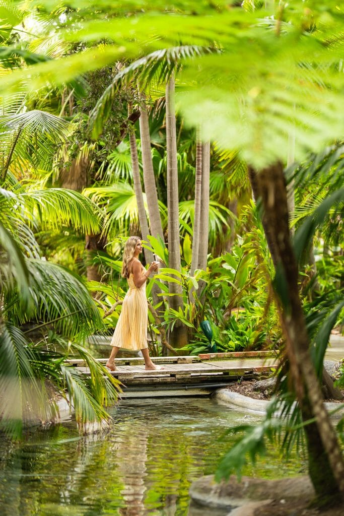 Woman in yellow dress walking on a wooden bridge over a small stream through a lush tropical forest at Bahia Resort Gardens