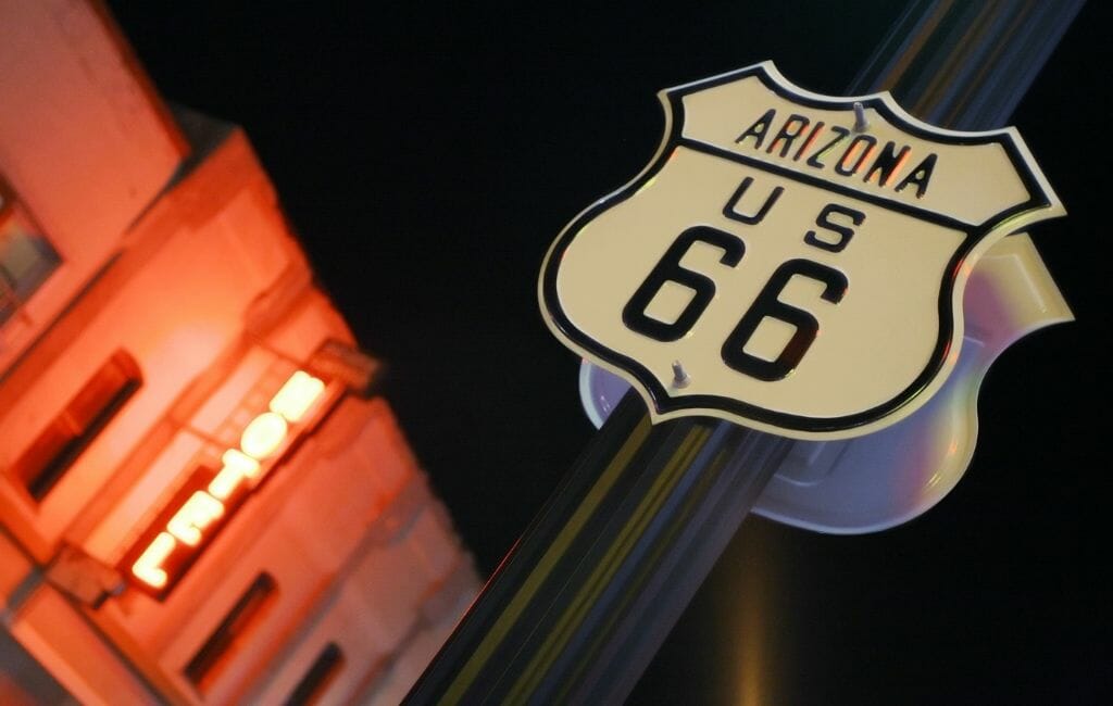 Historic Route 66 Sign in the foreground, dark sky in the background behind it, a blurry building lid up by red neon sign reading "hotel" on the left side of the image