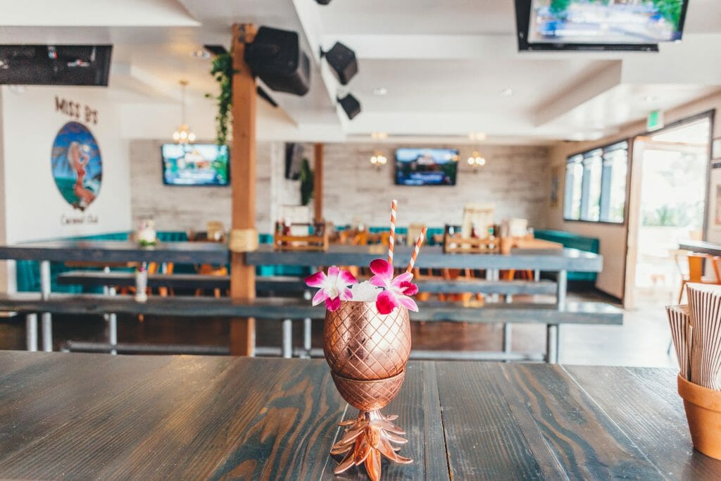foreground: Wood table with pineapple-shaped copper cocktail glass decorated with pink flowers, background empty bar