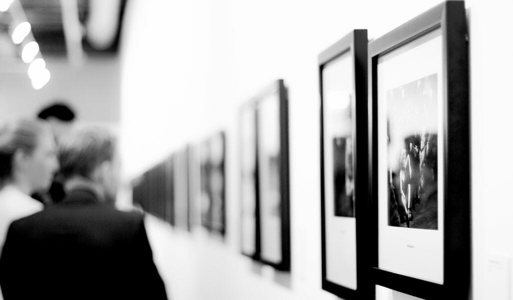 Black and white photo of a photography exhibition with black frames on the wall to the right and people out of focus in the distance to the left