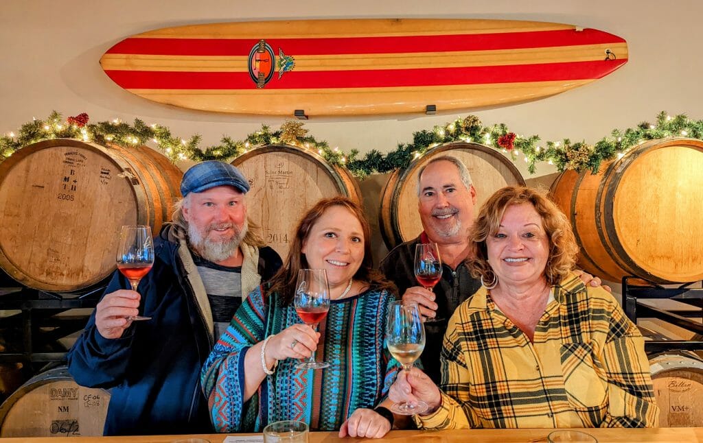 2 couples posing with wine glasses in front of wine barrels and a surfboard on the wall at Frisby Cellars Dana Point Harbor