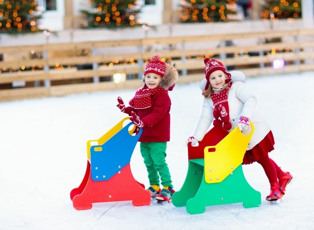 2 young girls dressed in red and white Winter dresses ice skating with  multicolored wood ice skate balancing devices