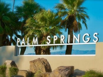 letter sign on top of tan colored wall spelling Palm Springs - Palm trees in the the background and blue sky