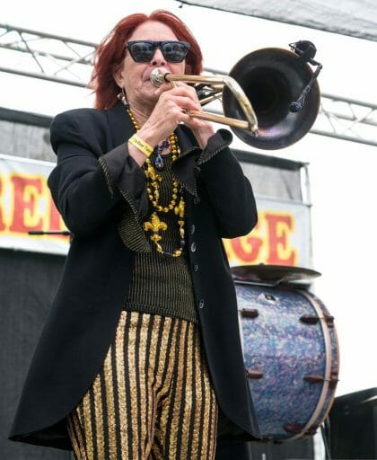 Woman with red hair and dark sunglasses, yellow black pants, black top and blazer playing trumpet