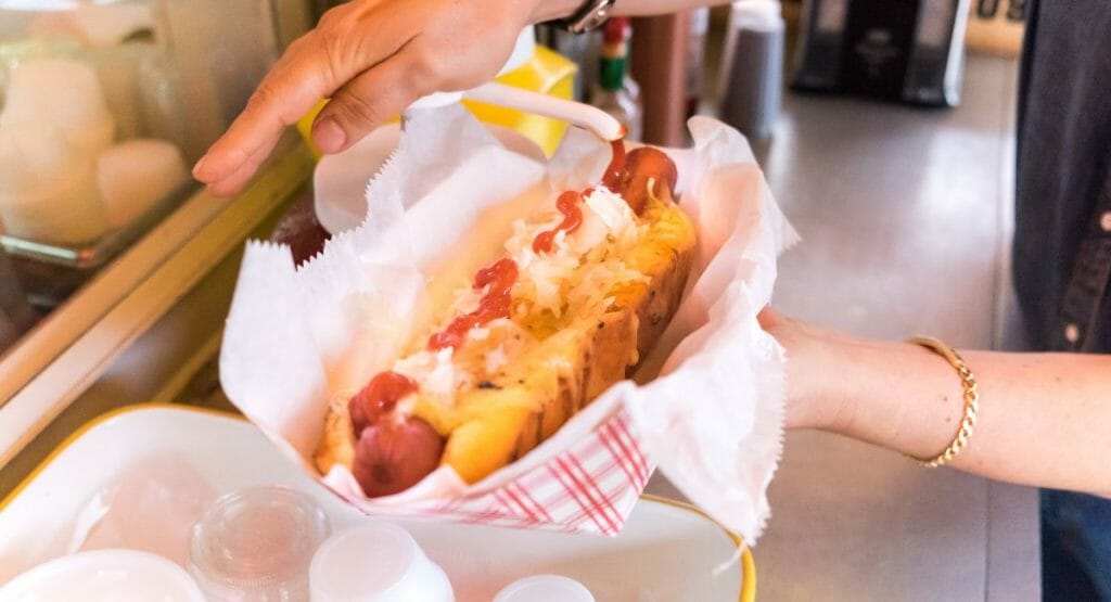 hands holding a hot dog with onions and ketchup in a paper container