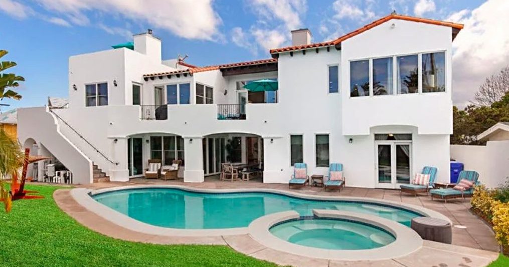 Spanish-style mansion with large pool in background surrounded by lounge chairs and green grass. Luxury vacation rentals in Coronado San Diego.