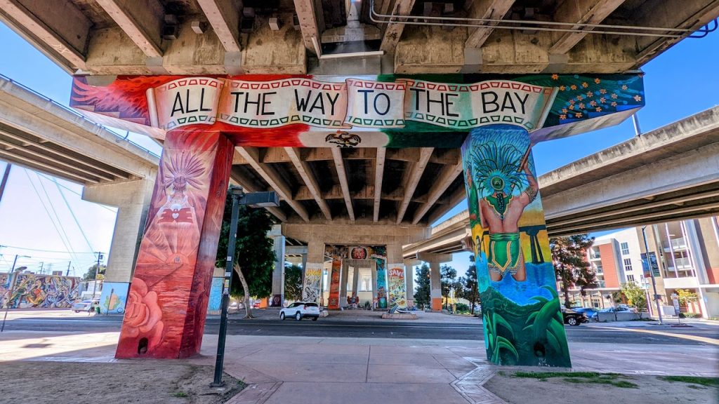 Large concrete beams painted in huge, colorful mural reading "all the way to the bay" on highway underpass. Chicano Park murals, Barrio Logan.