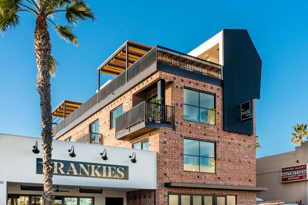 Three story brick building with balconies and rooftop deck overlooking palm trees. The Brick Hotel Oceanside.