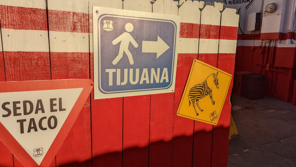 Red and white fence with signs showing TJ zonkey, stick figure with arrow towards Tijuana etc. 
