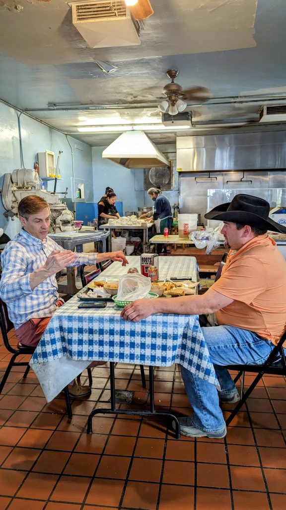 two men sitting on table in the front, women making tortillas in the back