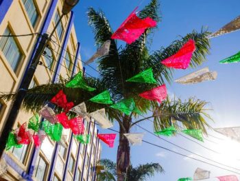 Red, green, and white paper flags with decorative cut outs, stringed above outdoor courtyard with building and palm tree in background on sunny day. Tijuana, Mexico Plaza Santa Cecelia. Tijuana tour from San Diego.