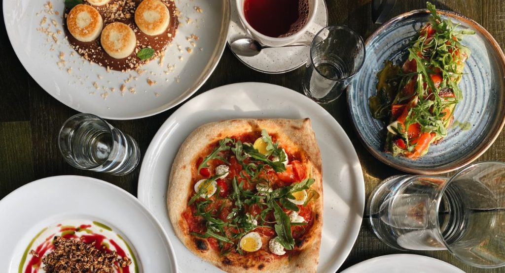 Photo of pizza and Italian food on wooden table surrounded by glassware at restaurant. Food&nbsp;tours San Diego.