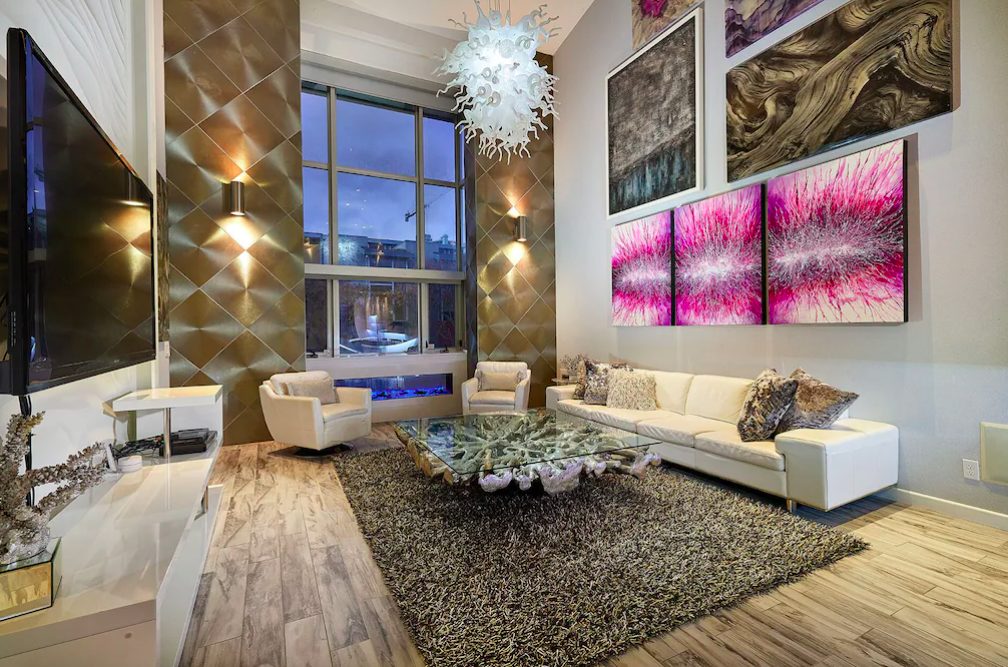 Living room with tall ceilings, floor to ceiling windows, colorful paintings on wall, and modern eccentric furnishings.