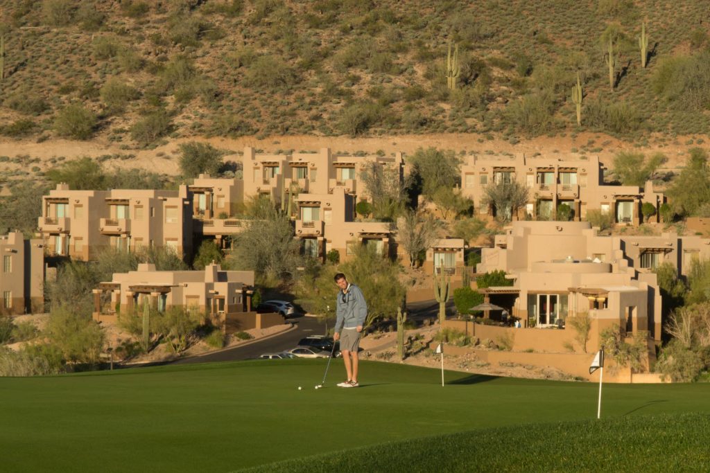 man playing golf in the foreground, sand colored resort Inn at Eagle Mountain in the background
