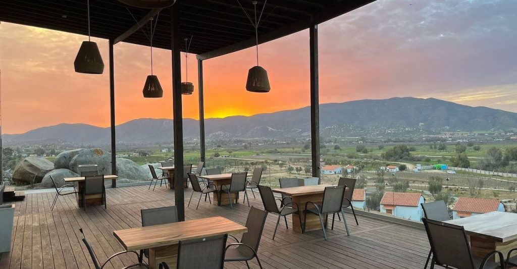 Wooden patio with restaurant table seating, patio is covered but completely open on sides with views of mountains in distance and desert valley below during bright orange sunset. Montevalle Health and Wellness Resort dining area.