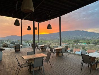Wooden patio with restaurant table seating, patio is covered but completely open on sides with views of mountains in distance and desert valley below during bright orange sunset. Montevalle Health and Wellness Resort dining area.