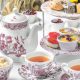 Fine China tea set with red florals and illustrations, matching tiered tray has finger foods and pastries on round table with white table cloth. High Tea San Diego.