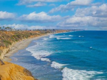 Blue water of the Pacific Ocean breaking on white sand beach next to sandy beach cliffs lined with palm trees on sunny day. North Ponto Beach, Carlsbad Beaches.