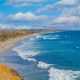 Blue water of the Pacific Ocean breaking on white sand beach next to sandy beach cliffs lined with palm trees on sunny day. North Ponto Beach, Carlsbad Beaches.