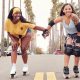 Two women holding hands while roller skating on street lined with cars and palm trees on hazy summer day. Roller Skating San Diego.