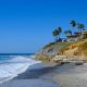 Waves breaking on sandy beach next to hill with large homes and palm trees on sunny summer day. Terramar Beach, Carlsbad Beaches.