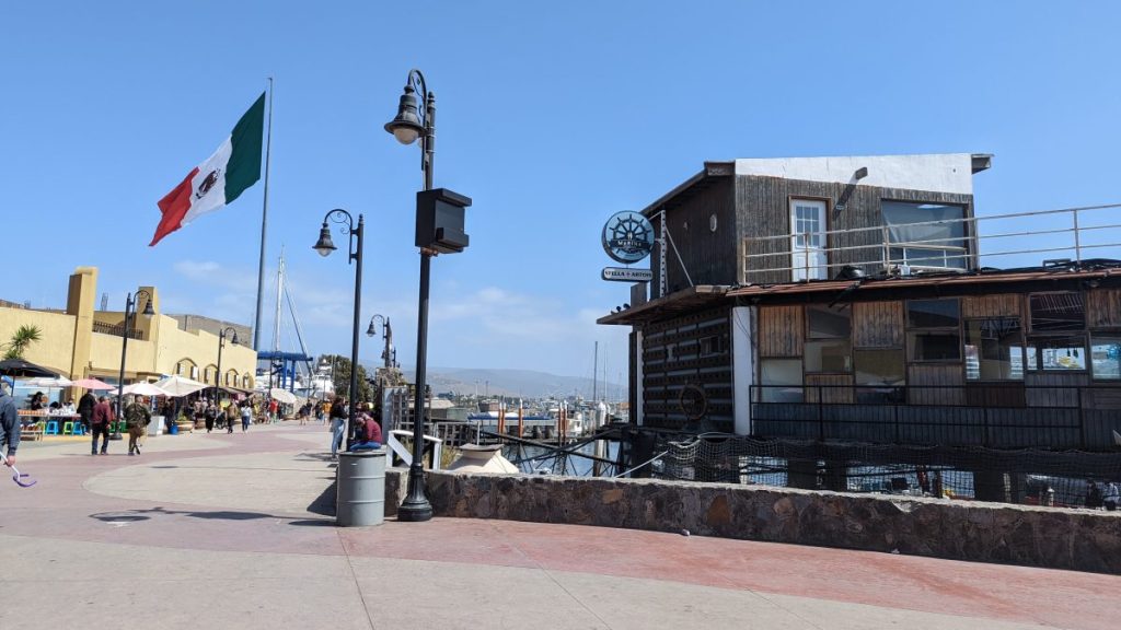 Promenade boardwalk lined with overwater buildings and shops, people walking along sidewalk with marina, mountains, and huge Mexico flag in distance on sunny day. Malecon, Things to do in Ensenada.
