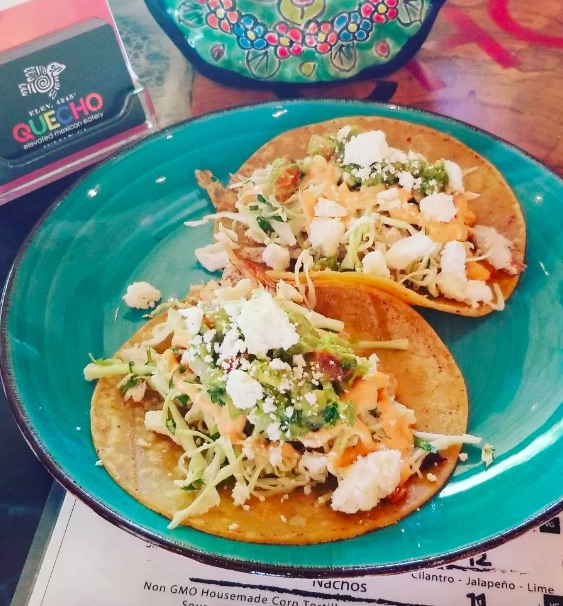 Two tacos on corn tortillas garnished with veggies and cheese on colorful, teal ceramic plate with "QUECHO" logo in background. Quecho Elevated Mexican Eatery, Julian Restaurants.