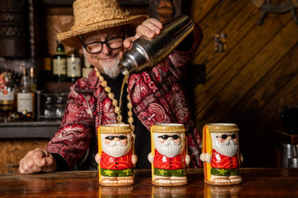 Man with straw hat pouring three cocktails in a santa mug - Tiki Christmas bars in San Diego
