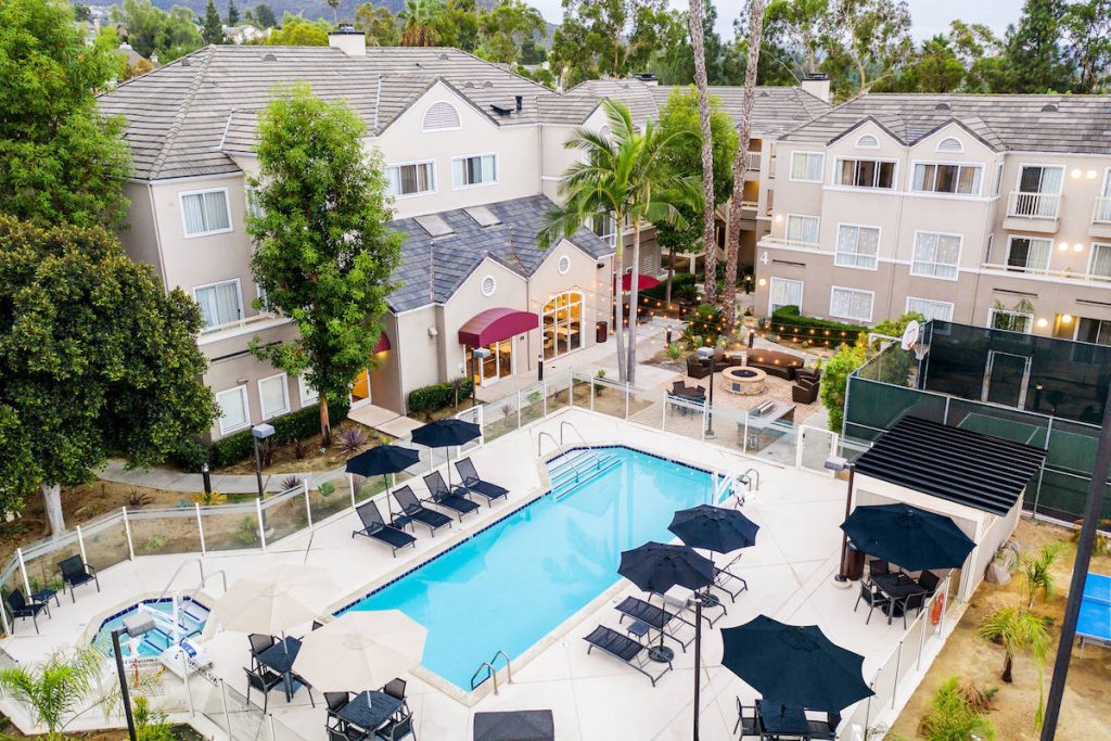 Aerial shot of hotel courtyard, outdoor pool, and tennis court surrounded by palm trees. Es Suites Carmel Mountain, hotel with balcony San Diego.