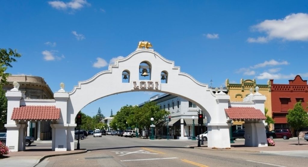 Large Spanish-style arch reading "Lodi" at entrance of small town on sunny day. Lodi, California wine regions.