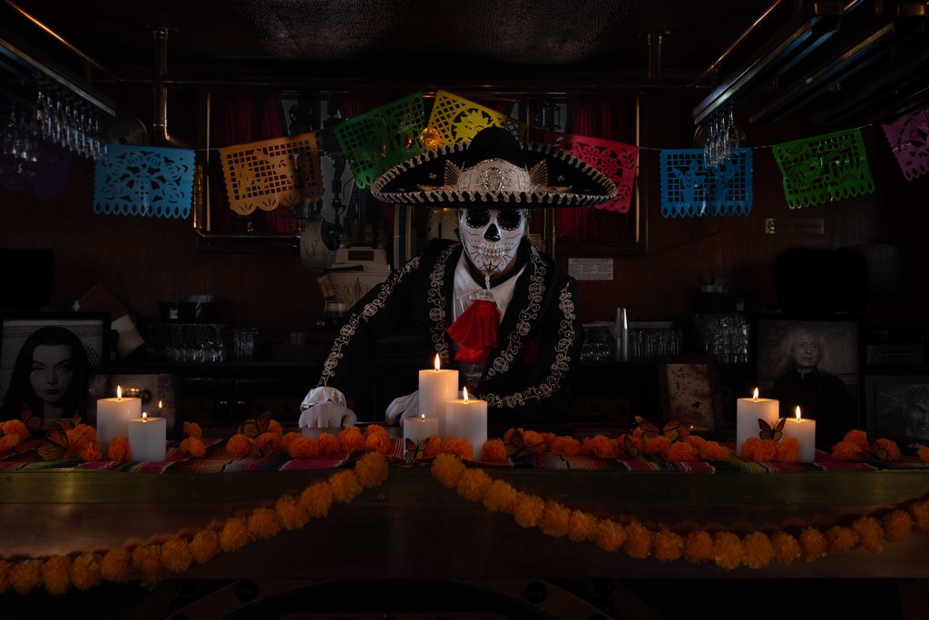 bartender with sugar skull makeup and sombrero behind a candle-lid bar